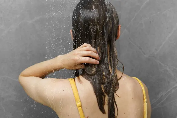 A young woman washes her wet hair with shampoo under a stream of water in the shower on a tile background. The concept of daily hygiene and self-care, beauty cosmetics