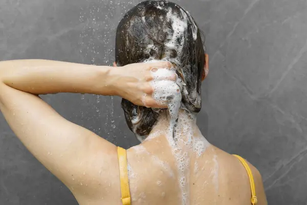 A woman washes her foamy hair with shampoo in the shower under water. The concept of scalp and hair care using natural balms, masks and shampoo