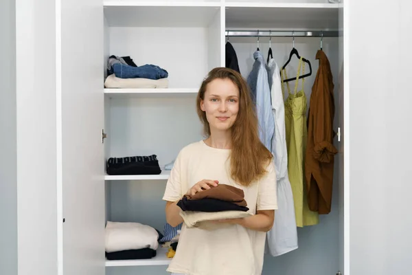 A young woman holds a stack of neatly folded clothing against a white closet with things. Personal stylist and buyer service concept, room cleaning, seasonal wardrobe change