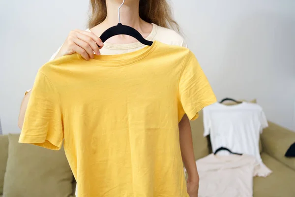 A young woman is holding a yellow mockup t-shirt on a hanger in front of her, choosing a look. Concept of seasonal wardrobe, shopping for new things. Image for your design