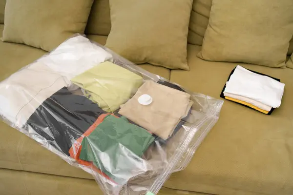 A transparent sealed bag filled with folded clothes lies on a green sofa. Concept of seasonal storage of clothes under vacuum to save space