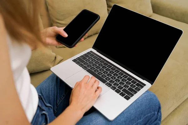 Young woman holding laptop and phone with black mockup screen sitting on couch. background of empty space for your content or advertising text.