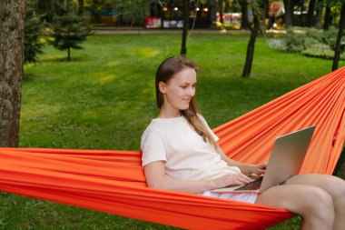 Genç bir kadın dizüstü bilgisayarla çalışırken ağaçların arasında turuncu bir hamakta otururken gülümsüyor. Uzaktan çalışma ve doğada eğlence kavramı.