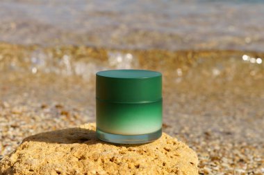 Kireç taşının üzerindeki bir kavanoz kremanın yeşil modeli güneş tarafından aydınlatılan deniz suyu ve dalgaların arka planına karşı. Güzellik ürünleri kavramı