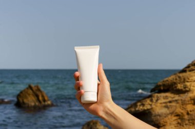 Deniz suyu ve taşlardan oluşan bir arka planda beyaz bir krema tüpü tutan bir kadının yakın çekimi. Güzellik ve estetiğin doğal ürünleri kavramı.