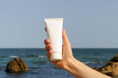 Deniz suyu ve plajın arka planında beyaz bir tüp vücut veya yüz kremi tutan bir kadın elinin yakın çekimi. Güzellik ve cilt bakım ürünleri kavramı