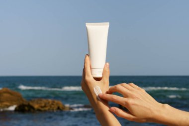 Kadın el beyaz bir krem tüpünü tutuyor ve plajın ve denizin arka planındaki deriye bir örnek uyguluyor. Cildi beslemek ve nemlendirmek için doğal kozmetik konsepti