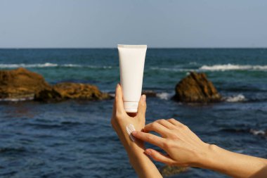 Kadın eli, beyaz, boş bir yüz veya vücut kremi tüpünü tutuyor ve deniz suyu ve plajın arka planında deri örneklerine bulaştırıyor. Doğal güzellik ürünleri kavramı