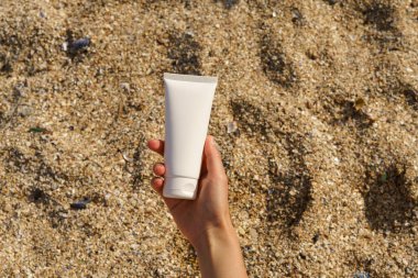 Bir kadının eli kumsaldaki bej kumun arka planında beyaz bir krem tüpünü tutuyor. Doğal kozmetik ürünleri, güzellik ürünleri kavramı