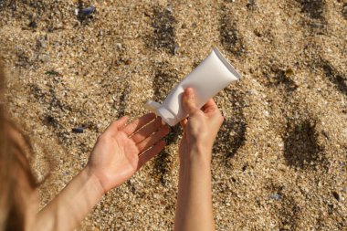 Kadın elleri kumsalın arka planında beyaz tüp modelinin cildine güneş kremi sürerler. Deniz özleri ile doğal kozmetik kavramı, cilt nemlendirici, yaz güzellik ürünleri