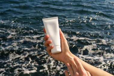 Dişi el, deniz suyunun arka planında beyaz bir krem tüpü tutuyor ve cilde bir örnek uyguluyor. Güneş koruması ve cilt suyu için güzellik ürünleri kavramı