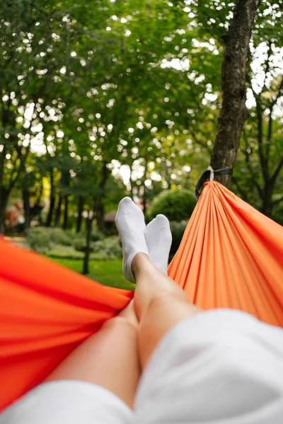 Vertical shot of a woman\'s leg in white socks resting on an orange hammock hanging from a tree. The concept of recovery in the midst of nature