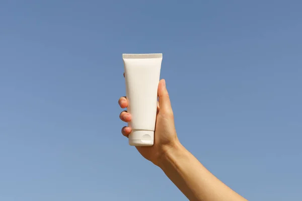 Female hand holding a white mockup tube of face or body cream on a blue sky background. The concept of natural cosmetics for nourishing and moisturizing the skin