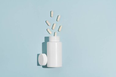 Mavi arka plan üzerine dökülen haplarla dolu beyaz maket kavanozu. Eczane, sağlık, ilaç, vitamin ve besin takviyesi kavramı.