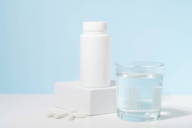 Bir kaidede beyaz bir hap kavanozu ve mavi arka planda bir bardak su. İlaç, eczane, güzellik ve sağlık vitaminleri, beslenme takviyeleri ve hastalıkların tedavisi kavramı.