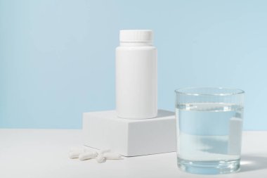 Kaidedeki bir kavanoz hapın beyaz maketi ve mavi arka plandaki bir bardak su. Eczane, vitamin, beslenme malzemeleri, sağlık, ilaç ve tedavi kavramı.