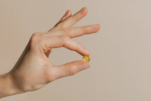 Female hand holding yellow transparent pill capsule with fingers on beige isolated background. Concept of pharmacy, depression treatment, vitamins and health improvement.