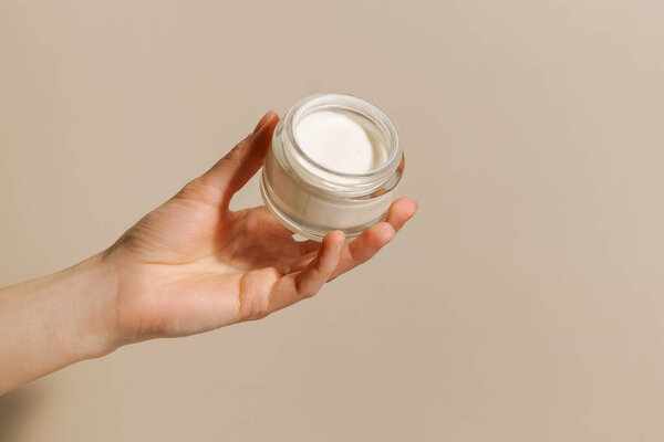 A female hand holds a glass jar with white face cream on a beige background. Concept of skin care, aesthetics, beauty products. Image for your design.