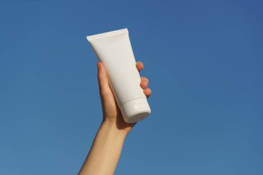 Mavi bir arka planda beyaz bir vücut tüpü ya da yüz kremi tutan kadın eli. Güneş koruması, nemlendirici ve besin kaynağı olan kozmetik kavramı, güzellik ürünleri.