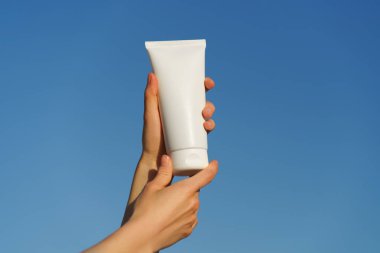 Mavi bir arka planda beyaz bir krem tüpünü tutan kadın eller. Vücut ya da yüz için doğal kozmetik kavramı, günlük bakım, spf ile korunma.