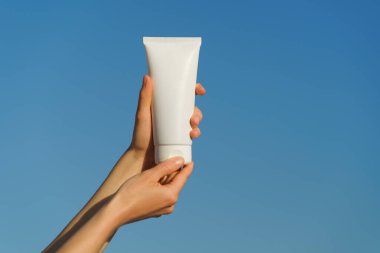 Kadın elleri mavi gökyüzü arka planında beyaz krema tüpünü açıyor. Güzellik kavramı, cildi nemlendirme, vücut bakımı, güneşten koruma, güneş kremi..