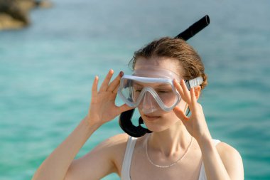 Beyaz mayo giymiş genç bir kadın, yüzünde şeffaf bir maske ve denizin arka planına dayanmış bir tüp tutuyor. Yaz eğlencesi ve eğlence konsepti, balık izleme, su dalışı ve şnorkelle yüzme.