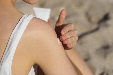 Genç bir kadın deniz kenarındaki kumun arka planında cildine güneş kremi sürüyor. Plajda yaz tatili kavramı, ultraviyole ışınlardan korunma, bronzlaşma.