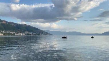 Deniz kıyısında dağlar, dalgalanan su ve suya yansıyan kalın bulutlar.