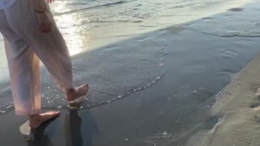 Islak kum ve deniz dalgaları üzerinde yürüyen genç bir kadının bacakları. Gerçek bir insanın yaşam tarzı, tatil, ilham, yalnız zaman.