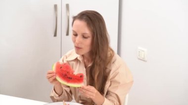 Bej gömlekli genç bir kadının, öğle veya akşam yemeklerini evde yerken, bir parça karpuz yerken ve beyaz bir masada otururken görüntüsü. Ev yemeği kavramı, sağlıklı atıştırmalık.