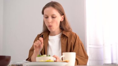 Kahverengi gömlekli genç bir kadın evde yemek yiyor. Tabaktan çatalla salata yiyor ve beyaz masada otururken çiğniyor. Ev yapımı yemek, sağlıklı beslenme, besleyici beslenme