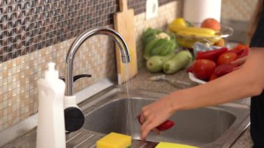 A young woman washes red pepper on the kitchen table in the sink under running water. The concept of home cooking, healthy eating, fresh vegetables