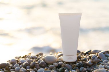 Çakıl taşları üzerindeki beyaz krem tüpü deniz zemininde batan güneş tarafından aydınlatılmıştır. Doğal kozmetik güzellik kavramı, cilt bakımı, güneş koruması