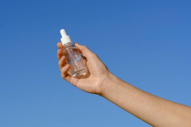 Mavi gökyüzü arka planında şeffaf cam bir kavanoz serum tutan kadın eli. Güzellik ürünleri, yüz bakımı, estetik kozmetoloji. Tasarımın için resim