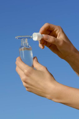 Mavi açık gökyüzü arka planında kadın ellerinin pipet dağıtıcıyı ve şeffaf cam serum kavanozunu tuttuğu dikey görüntü. Güzellik ürünleri, yüz bakımı, cilt nemlendirici.