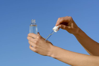 Kadın eli, pipet kutusuyla cilde serum uyguluyor ve mavi gökyüzü arka planında şeffaf cam kavanoz tutuyor. Güzellik ürünleri, yüz ve vücut bakımı, kozmetoloji prosedürleri ve spa