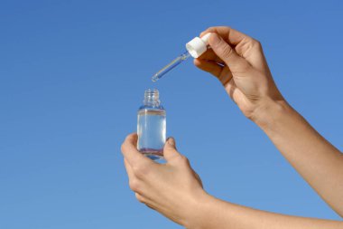 Mavi gökyüzü arka planında pipet makinesi ve şeffaf cam bir serum kavanozu tutan kadın eller. Cilt bakımı konsepti, reklam ve kozmetik ürünlerin sunumu