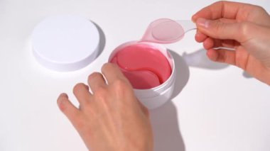 Kadın elleri spatulayla kavanozdan pembe bir yama çıkarır ve elinin üstüne beyaz masa arkasına koyar. Gözlerin altındaki güzellik ürünleri cildi yumuşatmak ve nemlendirmek için kullanılır..