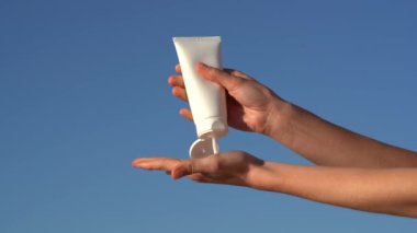 Kadın elleri, mavi gökyüzü arka planına karşı beyaz bir model tüpten avucunun içine krem veya losyon sıkıyor. Güzellik ürünleri, güneş koruması ve cilt suyu kavramı.