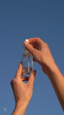 Bir kadın elinin cam bir serum kavanozunu açtığı ve mavi gökyüzü arka planındaki deriye pipet kutusundan uyguladığı dikey video. Güzellik ürünleri kavramı, nemlendirici ve besleyici yüz.