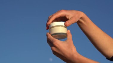 Kadın elleri cam bir krem kavanozunu açar ve kremi mavi gökyüzü arka planına sürterek cildine sürer. Güzellik ve vücut bakım ürünleri kavramı, doğal kozmetik ürünleri, güzellik ürünleri.
