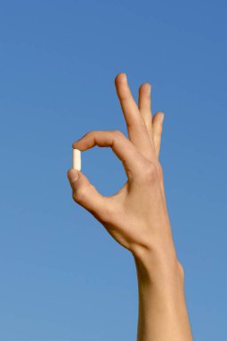 Mavi arka planda parmaklarıyla beyaz bir hapı tutan bir kadının dikey görüntüsü. Eczane ve ilaç konsepti, doğal sağlık hizmetleri, vitaminler ve takviyeler