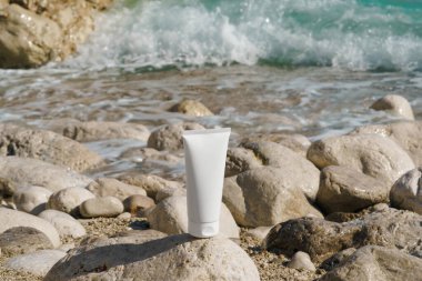 Sahildeki deniz suyunun arka planına karşı çakıl taşı taşının üzerindeki bir tüp kremin beyaz modeli. Doğal vücut bakımı kavramı, güzellik ürünleri, estetik..