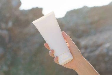 Bir kadının eli kayanın arka planında güneş tarafından aydınlatılmış beyaz bir krem tüpünü tutuyor. Güzellik ve cilt bakım ürünleri kavramı, nemlendirici, güneş koruması. Tasarımın için resim.