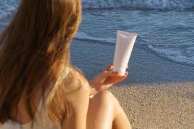 Seçici odaklı, dalgalı saçlı, kumsaldaki deniz dalgalarının arka planında beyaz bir krem tüpü tutan genç kadın. Vücut bakımı, güneş kremi, güzellik ürünleri için kozmetik konsepti.