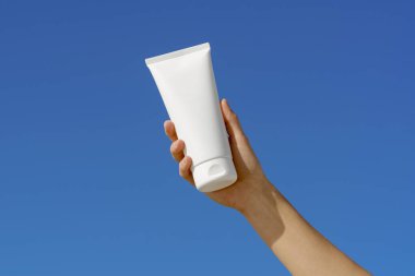 Mavi arka planda beyaz vücut ya da yüz kremi tüpü tutan bir kadın el. Güzellik ürünleri kavramı, güneş kremi, deri gıdası ve hidrasyon.