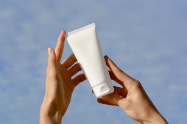 Mavi arka planda beyaz, boş bir krem tüpü ya da losyon tutan kadın eller. Vücut ve yüz bakımı için güzellik ürünleri kavramı, SPF ile korunma