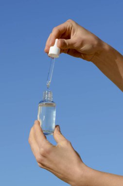 Mavi arka planda pipeti olan şeffaf cam bir serum kavanozunu tutan bir kadının dikey görüntüsü. Estetik ve güzellik ürünleri kavramı. Tasarımın için resim