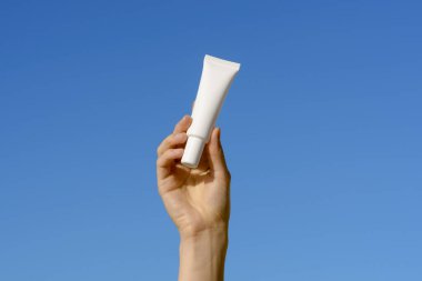Mavi gökyüzü arka planında beyaz, ince bir krem tüpü ya da serum tutan kadın eli. Yüz ve göz altı için nemlendirici kozmetik kavramı, güzellik bakımı