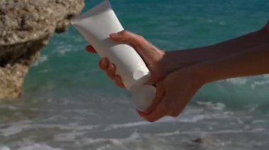 Genç bir kadın beyaz bir model tüp açar ve elindeki kremi deniz dalgalarının arka planına köpükle yapıştırır. Güneş kremi, cilt bakımı, güzellik ürünleri kavramı